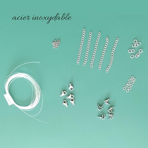 Könnte beinhalten: Eine Auswahl an Schmuckherstellungszubehör, darunter silberfarbene Kettenglieder, Verschlüsse, Sprungringe und kleine Perlen, angeordnet auf einer türkisfarbenen Oberfläche. Der Satz "acier inoxydable" steht in schwarzer Schrift oben links.