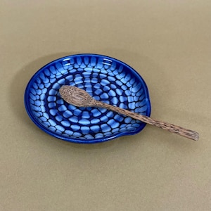 May include: A round ceramic spoon rest featuring a deep blue and white honeycomb-like pattern with a glossy finish. A small, light brown wooden spoon with a visible dark grain texture rests diagonally across the center of the dish.