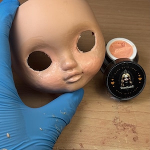 May include: A doll head with open eye sockets and painted facial features. Two small jars of peach-colored makeup are next to the doll head. One jar has a white lid, and the other has a black lid with the text "Domiladoll".