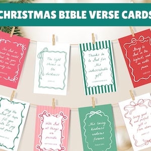 May include: Eight rectangular Christmas Bible verse cards hanging on a string with wooden clips. The cards feature red, white, green, and pink backgrounds with decorative borders and handwritten-style text containing various religious verses.