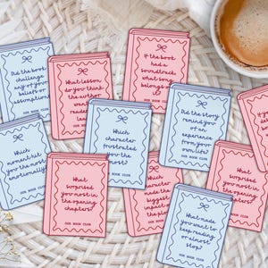 Op de afbeelding: Een verzameling rechthoekige, boekvormige kaarten in lichtblauw en roze, elk voorzien van een decoratieve geschulpte rand, een klein strikicoontje en gedrukte discussievragen voor een boekenclub, met de tekst Our Book Club onderaan.