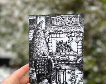 May include: Black and white Christmas card with a detailed illustration of a fireplace, Christmas tree, and a cozy armchair. The card features the text "Warmest Wishes" in a decorative banner. The card is held in a hand against a blurred outdoor background.
