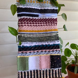 May include: A colorful woven wall hanging with a variety of textures and patterns. The hanging is made with different colored yarns and has a fringe at the bottom. The hanging is displayed against a white wall with a plant in a terracotta pot.