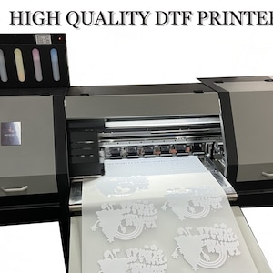 Pu&ograve; includere: Una stampante DTF di alta qualit&agrave; con un foglio di tessuto bianco che viene stampato con un design. La stampante ha un pannello di controllo con pulsanti e uno schermo di visualizzazione. La stampante &egrave; di colore nero e grigio.