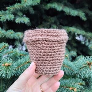 Può includere: Un piccolo vaso marrone all'uncinetto con una superficie strutturata. Il vaso è cilindrico con un bordo leggermente svasato. Lo sfondo è una vista sfocata di aghi di pino verdi.