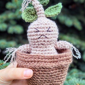 Può includere: Una pianta di Mandragora all'uncinetto in un vaso marrone. La pianta ha un corpo beige, foglie verdi e capelli bianchi filiformi. Il vaso è di una tonalità di marrone più scura. La Mandragora ha un'espressione scontrosa.