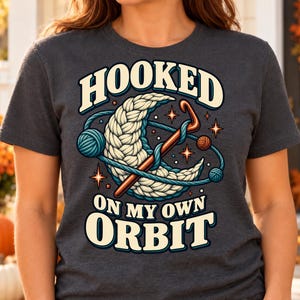 Könnte beinhalten: Ein dunkel anthrazitfarbenes T-Shirt mit einer Grafik aus einem weißen Garn-Halbmond, einer braunen Häkelnadel und kleinen Garnknäueln, die wie Planeten kreisen. Der Text HOOKED ON MY OWN ORBIT ist in fetten, cremefarbenen Blockbuchstaben gedruckt.