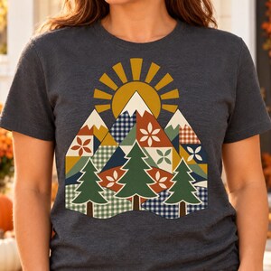 Puede incluir: Una camiseta de manga corta en gris jaspeado oscuro con un estampado gráfico de montañas estilizadas y tres pinos. El diseño de la montaña incorpora patrones geométricos, incluyendo cuadros vichy y motivos florales, bajo un sol amarillo.
