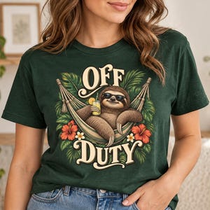 May include: A dark green short-sleeve t-shirt featuring a graphic of a sloth wearing sunglasses, relaxing in a hammock while holding a yellow drink. The text "OFF DUTY" is printed in a cream-colored font above and below the sloth, surrounded by tropical green leaves and red hibiscus flowers.