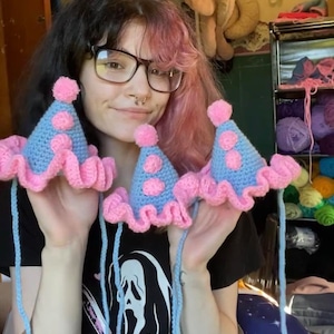 May include: Crocheted party hats in light blue with pink polka dots and a pink ruffled brim. Each hat has a pink pom-pom on top and long blue strings. The hats are handmade and appear to be for a pet or small child.