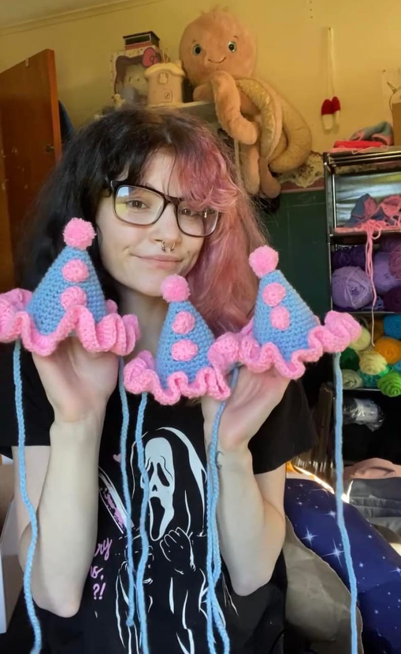 May include: Crocheted party hats in light blue with pink polka dots and a pink ruffled brim. Each hat has a pink pom-pom on top and long blue strings. The hats are handmade and appear to be for a pet or small child.