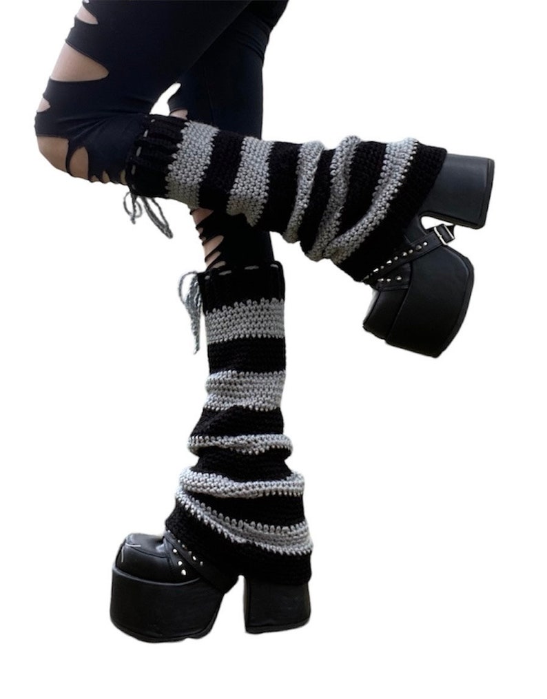 Pu&ograve; includere: Scaldamuscoli a righe nere e grigie. Gli scaldamuscoli sono realizzati in materiale a maglia e sono indossati con stivali con plateau neri. Gli stivali hanno fibbie e borchie. Gli scaldamuscoli sono un accessorio di moda.