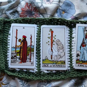 Può includere: Un mazzo di carte dei tarocchi con dorsi blu e tre carte esposte in un supporto all'uncinetto verde. Le carte mostrano illustrazioni di figure e simboli, tra cui le carte "Asso di Bastoni" e "Re di Spade".