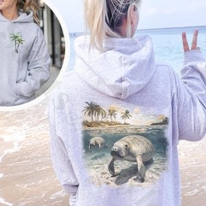 May include: Light gray hoodie with a manatee graphic on the back. The graphic depicts manatees in the ocean with palm trees and a beach scene. The front of the hoodie has a small palm tree design. The word "ash" is at the bottom.