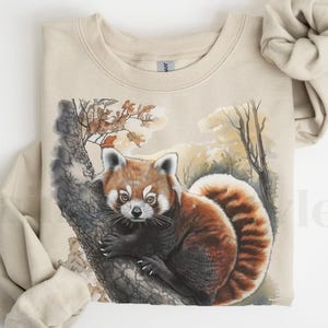 May include: A beige sweatshirt with a red panda illustration. The panda is sitting on a tree branch with leaves and a watercolor background. The sweatshirt has a crew neck and long sleeves.