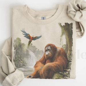Op de afbeelding: Een beige sweatshirt met een grafische print van een orang-oetan die in een jungle-omgeving zit met een blauwe en rode ara die erboven vliegt. Het sweatshirt heeft een ronde hals en lange mouwen.