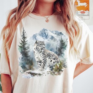 May include: A white t-shirt with a watercolor design of a snow leopard in a snowy mountain landscape. The shirt is ivory in color.