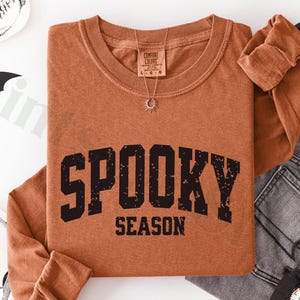 May include: A burnt orange long-sleeve shirt with the words "SPOOKY SEASON" in black block letters. The shirt is paired with gray jeans and Halloween-themed decorations, including a skull and bats.
