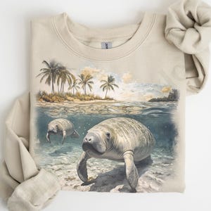 May include: A tan sweatshirt with a graphic print of two manatees swimming in the ocean. The print features palm trees and a sunset in the background.