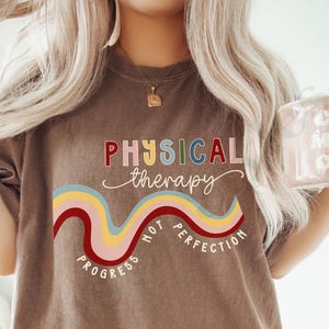 May include: An espresso-colored t-shirt featuring the text PHYSICAL THERAPY in multi-colored letters, a wavy line graphic in red, yellow, and blue, and the text PROGRESS NOT PERFECTION curved underneath the graphic in white lettering.