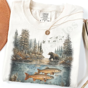 May include: Ivory-colored t-shirt with a nature scene featuring a bear in a river, fish, trees, and birds. The shirt has a Comfort Colors tag and a gold necklace. The word "Ivory" is printed in the upper right corner.