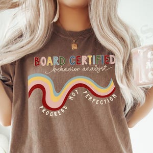 May include: Espresso-colored t-shirt with the text "BOARD CERTIFIED behavior analyst" above a wavy rainbow design. Below the design are the words "PROGRESS NOT PERFECTION". The shirt is a casual style.