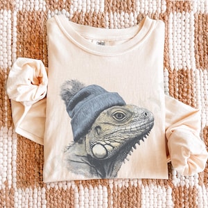 May include: Ivory long-sleeve shirt featuring a detailed illustration of an iguana wearing a blue beanie with a gray pom-pom. The shirt is made of a soft material and is laid flat on a patterned surface.