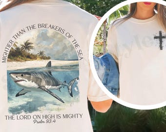 Puede incluir: Camiseta color marfil con una ilustración de tiburón y el texto "MIGHTIER THAN THE BREAKERS OF THE SEA" y "THE LORD ON HIGH IS MIGHTY Psalm 93:4". Una cruz negra está en la parte delantera.