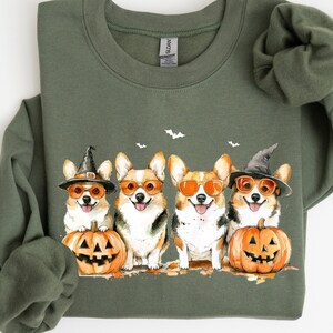 May include: A green sweatshirt with a Halloween-themed design featuring four corgis wearing witch hats and sunglasses, holding pumpkins. The corgis are surrounded by bats and fall leaves.