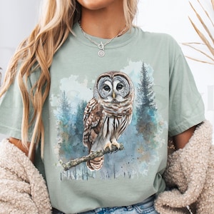 May include: A light green t-shirt with a watercolor illustration of an owl in a forest setting. The owl is brown and gray with large, round eyes. The text "Bay" is visible in the lower left corner of the image.