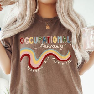 May include: A brown t-shirt with the text "Occupational Therapy" in colorful letters. The text is surrounded by a colorful wavy line with the words "Progress Not Perfection" underneath.