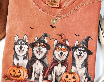 May include: Orange Comfort Colors t-shirt featuring four smiling huskies dressed in Halloween costumes, including witch hats and capes. Each dog is next to a carved pumpkin. A gold necklace and a brown hat are also visible.