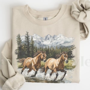 May include: A beige sweatshirt with a graphic print of two brown horses running through a stream in front of a mountain range. The mountains are covered in snow and the trees are green.