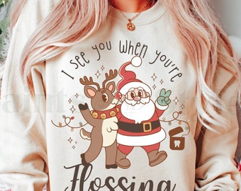May include: A beige sweatshirt with a Christmas-themed graphic featuring Santa Claus and a reindeer. The graphic says "I see you when you're flossing".