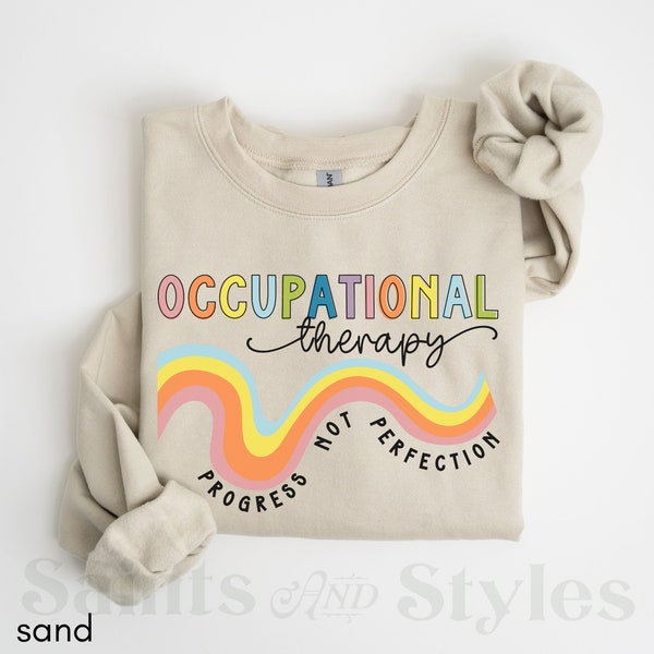 Può includere: Una felpa beige con il testo "Occupational therapy" in lettere arcobaleno. Il testo "Progress not perfection" è scritto in un arcobaleno sotto il testo principale.