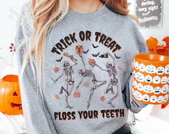 May include: A gray sweatshirt with the text "Trick or Treat Floss Your Teeth" in orange and black. The design features three skeletons holding Halloween themed items, including a toothbrush, toothpaste, and a bucket of candy.