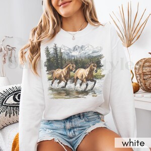 May include: White sweatshirt with a graphic of two brown horses running through a mountain stream. The mountains are snow-capped and the trees are green.