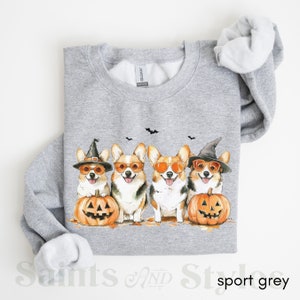 May include: A gray sweatshirt with a graphic of four corgis wearing witch hats and sunglasses, holding jack-o'-lanterns. The corgis are in a row, facing forward. The sweatshirt has a crew neck and long sleeves.