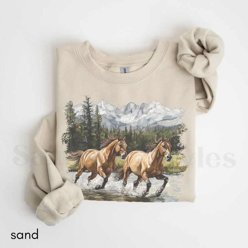 May include: A beige sweatshirt with a graphic print of two brown horses running through a stream in front of a mountain range. The mountains are covered in snow and the trees are green.