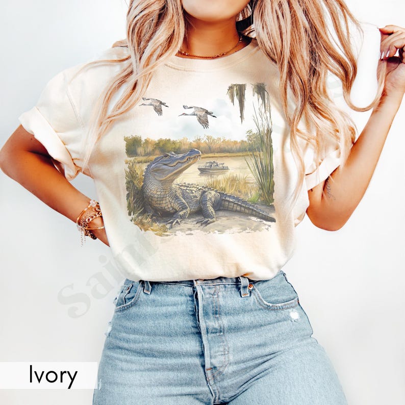 May include: Ivory-colored t-shirt featuring a detailed illustration of an alligator resting near a body of water, with two birds flying overhead. A boat is visible in the background. The word "Ivory" is printed at the bottom.