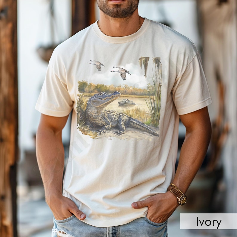May include: Ivory-colored t-shirt featuring a detailed illustration of an alligator resting near a body of water, with two birds flying overhead. The design includes a boat and marsh scenery. The shirt is a neutral color.
