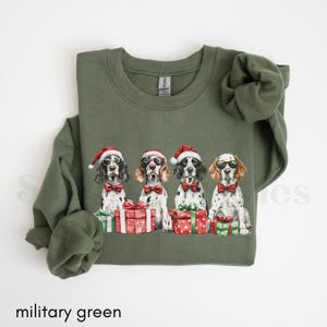 May include: A military green sweatshirt with a Christmas-themed design featuring four dogs wearing Santa hats and holding presents. The dogs are all wearing sunglasses and have a festive look.