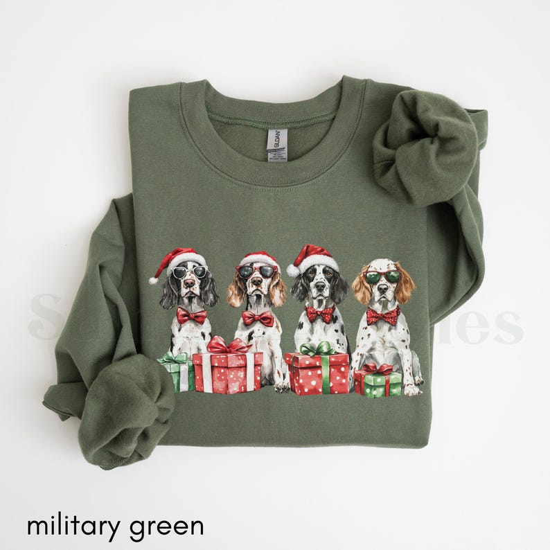 May include: A military green sweatshirt with a Christmas-themed design featuring four dogs wearing Santa hats and holding presents. The dogs are all wearing sunglasses and have a festive look.