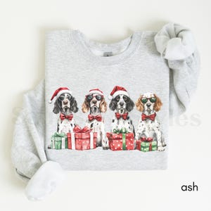 May include: A gray sweatshirt with a Christmas-themed design featuring four dogs wearing Santa hats and sunglasses. The dogs are sitting in front of presents with red and green wrapping paper.