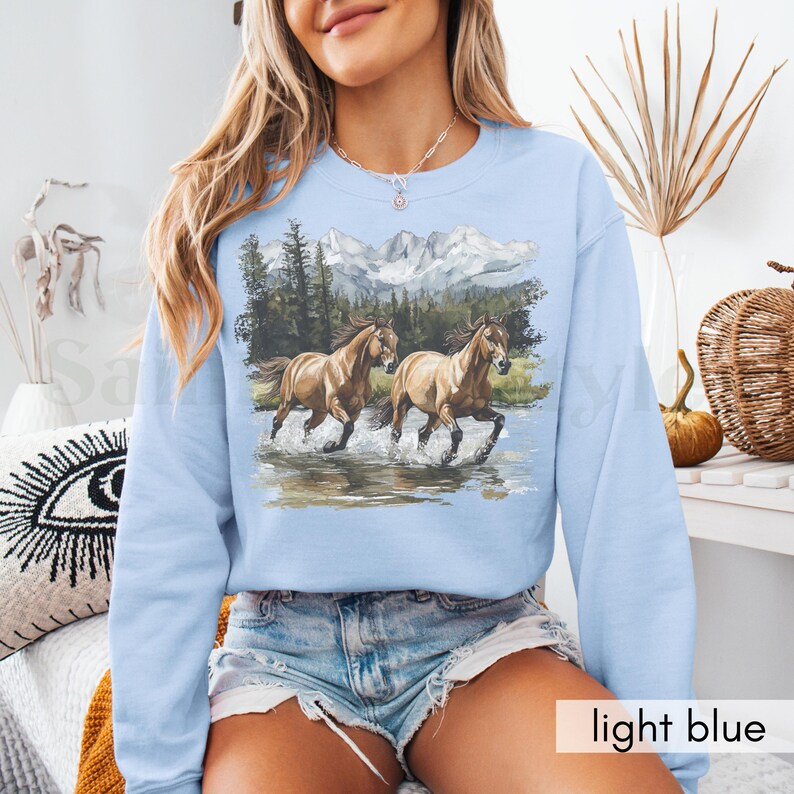 May include: A light blue sweatshirt with a graphic of two brown horses running through a mountain landscape. The horses are in a forest setting with trees and snow-capped mountains in the background.