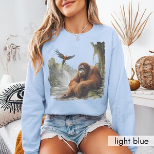 Op de afbeelding: Een lichtblauw sweatshirt met een grafische print van een orang-oetan die op een rots zit met een papegaai die erboven vliegt. De orang-oetan is bruin en de papegaai is rood, geel en blauw.