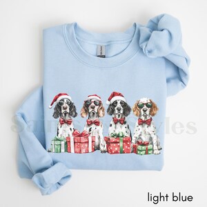 May include: Light blue sweatshirt with a Christmas-themed design featuring four dogs wearing Santa hats and holding presents. The dogs are all wearing bow ties and have a festive look.