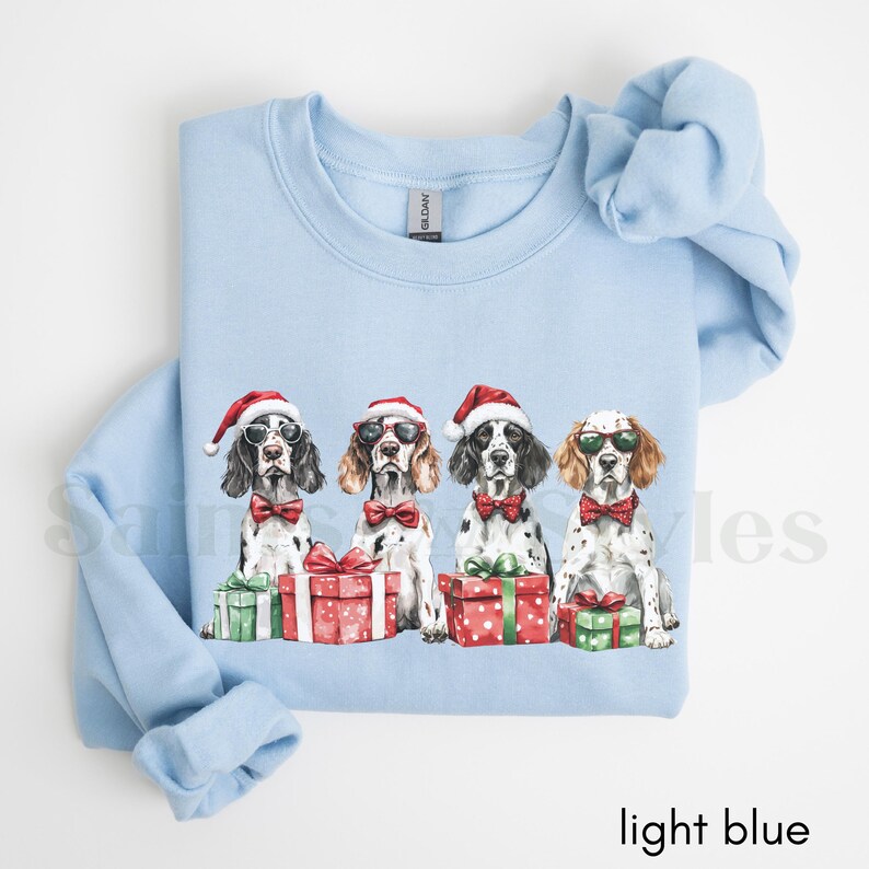 May include: Light blue sweatshirt with a Christmas-themed design featuring four dogs wearing Santa hats and holding presents. The dogs are all wearing bow ties and have a festive look.