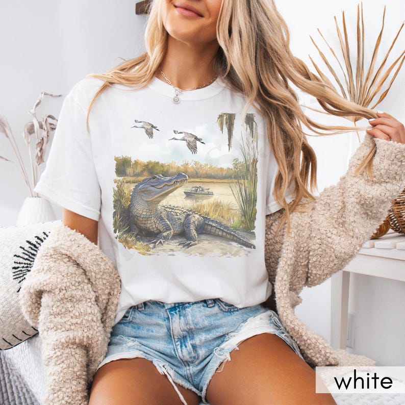 May include: White t-shirt featuring a watercolor illustration of an alligator resting on a riverbank, with two birds flying overhead. The scene includes a boat on the water and tall grass. The word "white" is printed at the bottom.
