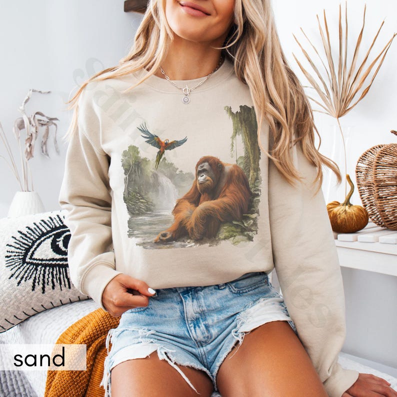 Op de afbeelding: Een beige sweatshirt met een grafische print van een orang-oetan die in een jungle-omgeving zit met een papegaai die erboven vliegt. Het sweatshirt heeft een ronde hals en lange mouwen.